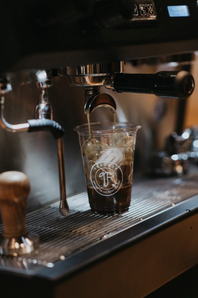 Close-up of iced coffee being brewed from an espresso machine into a plastic cup indoors.